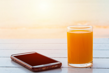 Glass of orange juice with the phone placed on a table by the sea.の写真素材