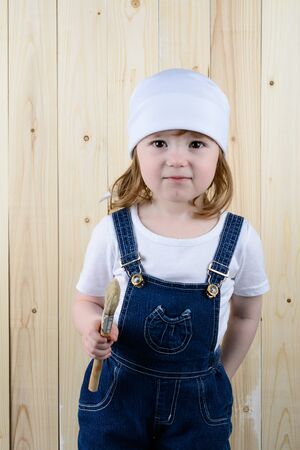 Little girl in the form of a painter with a brush in his hands posing on the background of wooden wallの写真素材