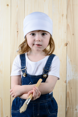 Little girl in the form of a painter with a brush in his hands posing on the background of wooden wallの写真素材