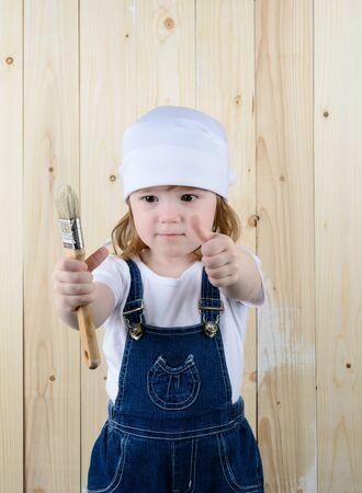 Little girl in the form of a painter with a brush in his hands posing on the background of wooden wallの写真素材