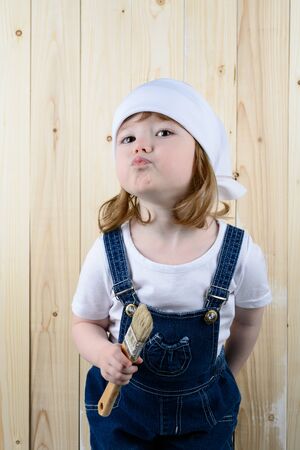Little girl in the form of a painter with a brush in his hands posing on the background of wooden wallの写真素材