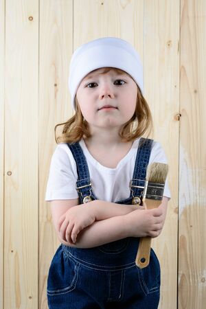 Little girl in the form of a painter with a brush in his hands posing on the background of wooden wallの写真素材
