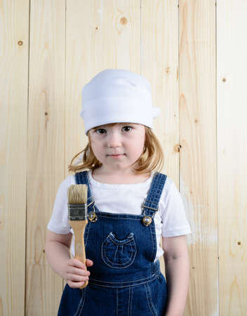 Little girl in the form of a painter with a brush in his hands posing on the background of wooden wallの写真素材