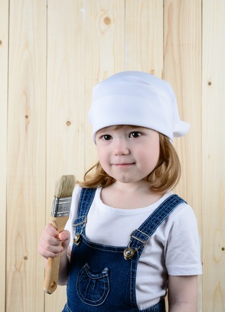 Little girl in the form of a painter with a brush in his hands posing on the background of wooden wallの写真素材
