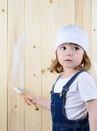 Little girl in the form of a painter paints a wooden wall in whiteの写真素材