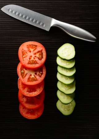 Cut tomato and cucumber with a knife lie on a dark tileの写真素材