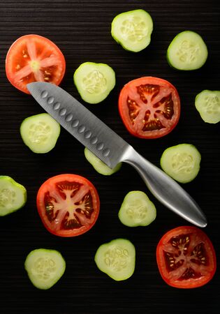 Cut tomato and cucumber with a knife lie on a dark tileの写真素材