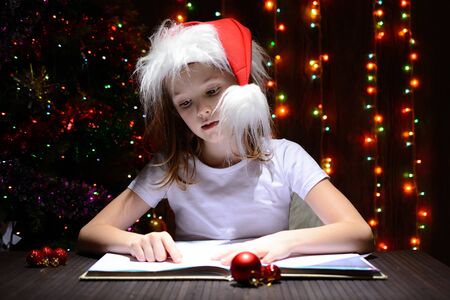 Girl in santa hat reading a book on a background of luminous garlandsの写真素材
