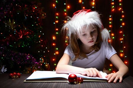 Girl in santa hat reading a book on a background of luminous garlandsの写真素材