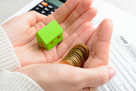 Coins and paper house in female hands and tax form on the tableの写真素材