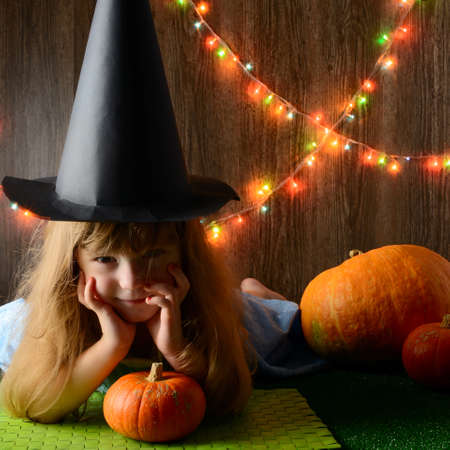 Young girl wearing Halloween hat holding pumpkin sitting against wall with colorful lampsの写真素材