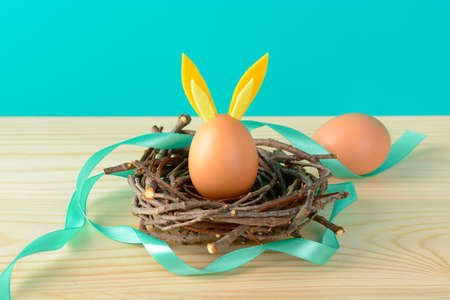Composition of chicken egg with rabbit ears and blue ribbon lying in twig nest on blue backgroundの写真素材