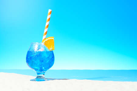 Refreshing blue drink with ice and a slice of orange in a glass on the sandy coastの写真素材