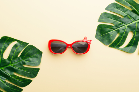 From above shot of green Monstera leaves surrounding red sunglasses on beige surface.の写真素材