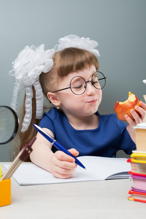 Female kid in glasses and white bows smiling with fruit and doing homework with blue penの写真素材