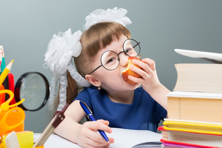 Female kid in glasses and white bows smiling with fruit and doing homework with blue penの写真素材