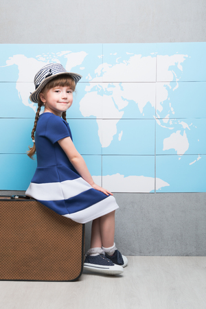 Cute female child smiling and looking at camera on baggage with countries on backgroundの写真素材