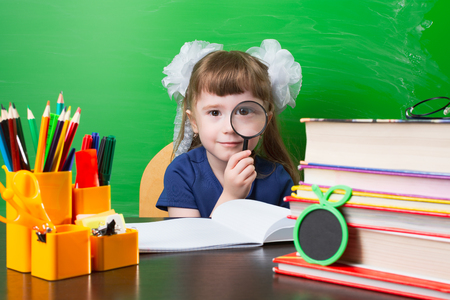 Little girl with tails watching glass at table with school supplies against green wallの写真素材