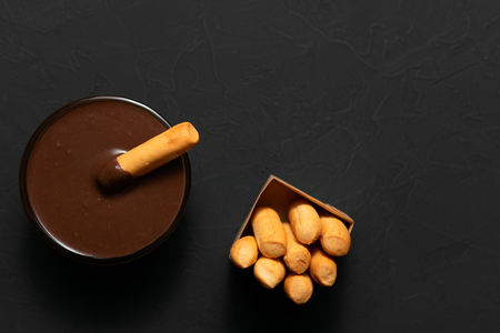 Top view of bowl of melted chocolate for dipping golden crispy breadsticks composed on black backgroundの写真素材