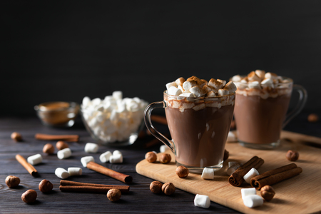 Transparent cups with hot chocolate and delicious sweet candies on chopping board near scattering cinnamon and nuts on black backgroundの写真素材