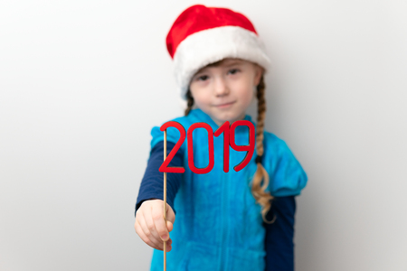 Closeup of red number 2019 in hands of little girl wearing Santa hat on white backgroundの写真素材