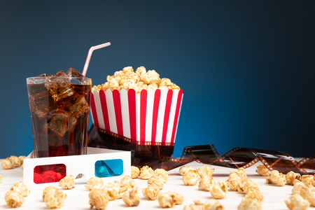 Cinema concept of 3D glasses, snacks scattering near paper cup, glass of soda with ice and film strip on white board on blue backgroundの写真素材