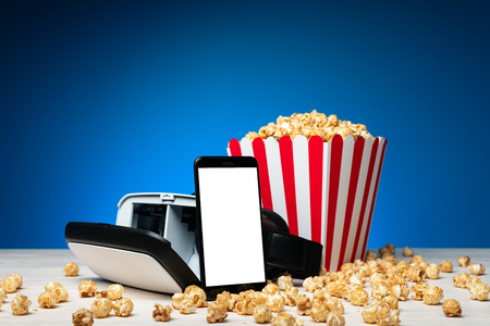 Arrangement of modern headset of virtual reality with smartphone and colorful bucket of popcorn on blue backgroundの写真素材