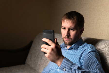 Adult male sitting on comfortable couch at home and browsing modern smartphoneの写真素材