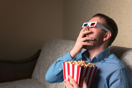 Handsome adult guy in 3D glasses covering mouth in shock while eating popcorn and watching movie on comfortable sofaの写真素材