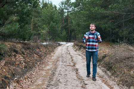 Handsome guy with backpack looking away while walking along dirty road in conifer forestの写真素材