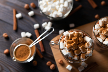 Top view mugs with hot chocolate and marshmallows on chopping board near scattering cinnamon, cocoa and nuts on black wood backgroundの写真素材