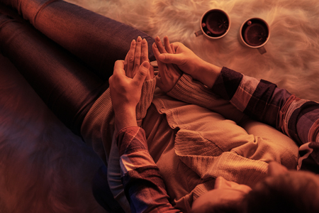 From above crop shot of embracing couple cuddling on furry carpet in light of fireplace having warming drinksの写真素材