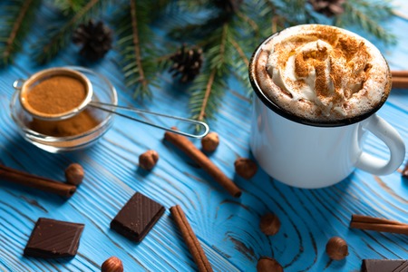 Mug with hot chocolate and whipped cream among cinnamon sticks, nuts on a blue wooden tableの写真素材