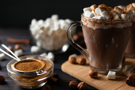 Transparent cups with hot chocolate and delicious sweet candies on chopping board near scattering cinnamon and nuts on black backgroundの写真素材
