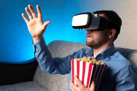 Adult guy with fresh popcorn wearing modern VR headset and gesturing with hand while sitting on comfortable couch at homeの写真素材