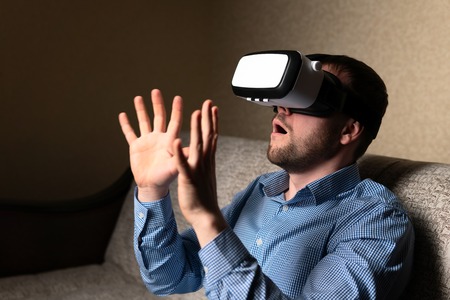 Adult male in VR glasses waving hand while sitting on comfortable sofa at homeの写真素材