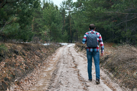 Back view of male with backpack walking along dirty countryside road while travelling through conifer forestの写真素材