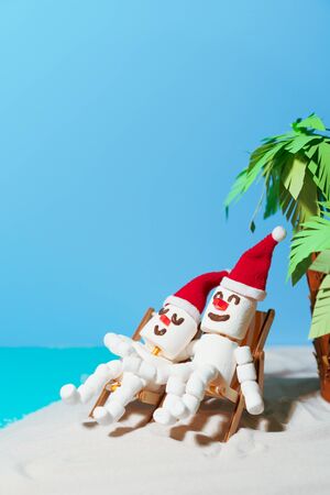 Two marshmallow snowmen in love are sitting on deckchairs on a sandy beach. Christmas holidays on the beach.の写真素材