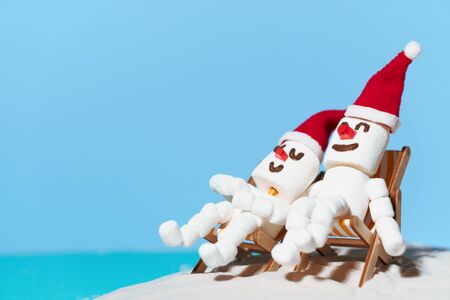 Two marshmallow snowmen in love are sitting on deckchairs on a sandy beach. Christmas holidays on the beach.の写真素材