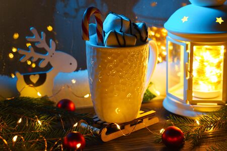 White mug with hot chocolate and marshmallows with Christmas decorations on the windowsill on a winter evening at the blue window.の写真素材