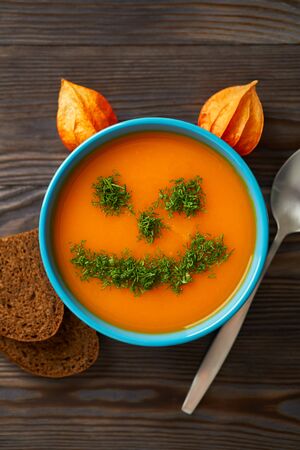 Autumn cream soup garnished with dill in the form of funny faces Halloween pumpkins. Healthy pumpkin and carrot soup puree in a blue bowl on a wooden background. Halloween holiday concept, top view.の写真素材