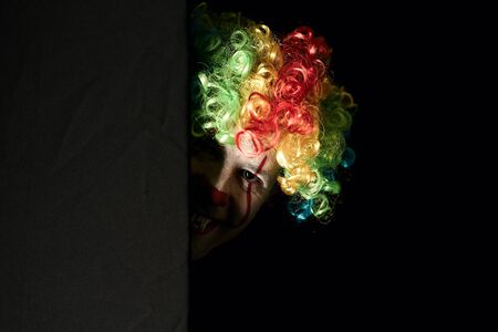 Close-up of a scary clown peeping around the corner of a black wall. He is wearing a colored wig and...