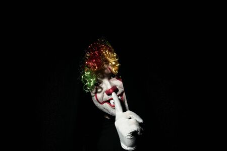 A terrible scary clown in a colored wig peeps out from behind black curtains. Shows a gesture of silence with a finger at the lips.の写真素材