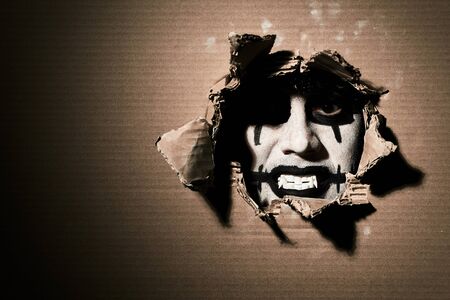 A creepy scary vampire clown looks through a torn cardboard. Halloween theme with blank space on the...