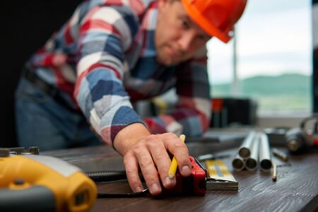 Young guy engineer in a plaid shirt holds out his hand with a pencil to the measuring tape. Focus on...