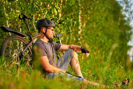 A cyclist sits on the grass and looks at the sunset. A man in a helmet next to a bicycle in nature.の写真素材