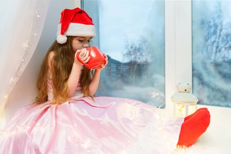 Cute little girl in a pink dress and santa hat sits on a windowsill with a red mug of hot chocolate in her hands.の写真素材