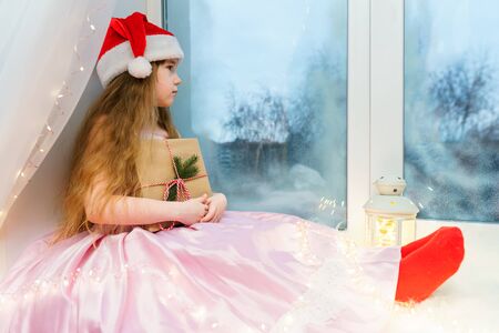 Charming little girl in a pink dress sits on a windowsill and holds a Christmas present in her hands, looking out the window.の写真素材