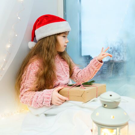 A charming little girl in a pink sweater with a gift in her hands sits on a windowsill and looks at the street through a frozen window.の写真素材