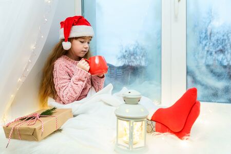 Cute little girl in a pink sweater and santa hat sits on a windowsill with a red mug of hot chocolate in her hands.の写真素材